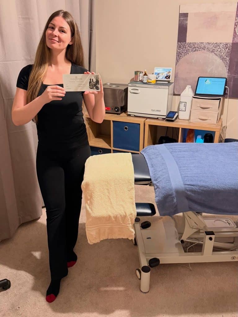 Canan Burden in her treatment room, standing beside the massage table and holding a gift voucher